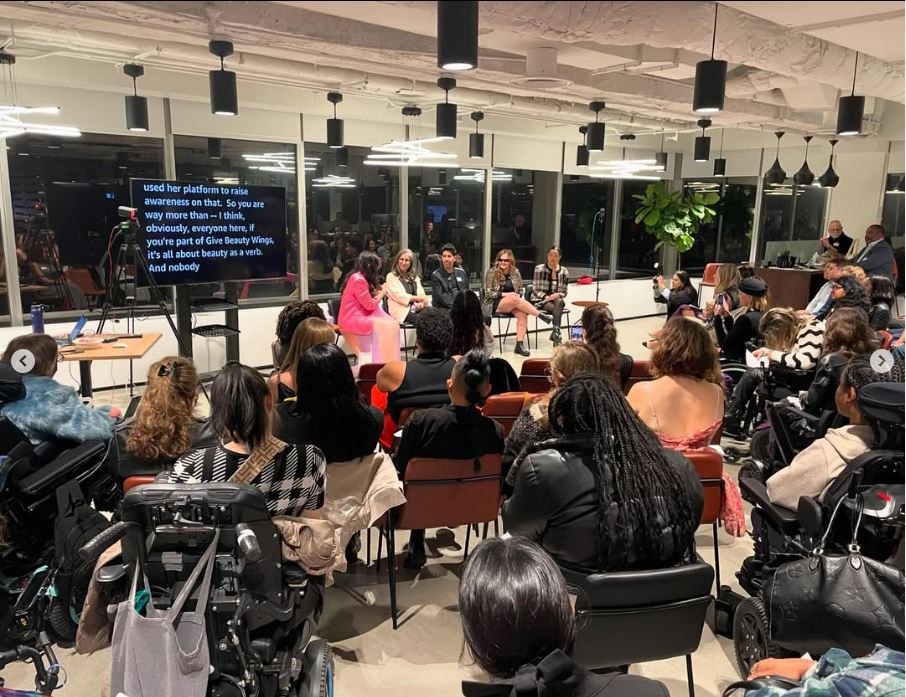 A panel discussion with 5 speakers sitting next to a CART live text monitor, with audiences sitting facing them, some on chairs, some using wheelchairs. 